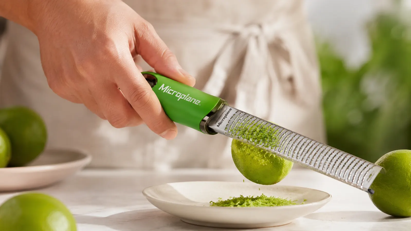 A hand running the Microplane zester over a lime, with fine curls of zest falling onto a small white plate